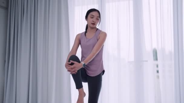 Μια Ασιάτισσα γυναίκα με στολή γυμναστικής τεντώνει με χάρη τους μυς της χρησιμοποιώντας γιόγκα ή στάσεις άσκησης σε ένα χαλάκι στο καθιστικό του σπιτιού της. Ιδανικό για fitness ή yoga προώθηση. - Πλάνα, βίντεο
