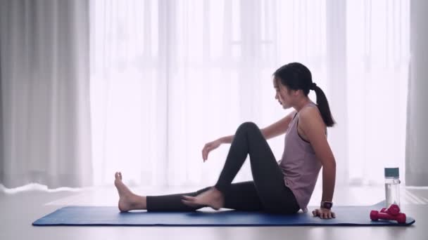 Uma mulher asiática em traje de treino graciosamente alonga seus músculos usando ioga ou poses de exercício em um tapete em suas casas sala de estar. Ideal para fitness ou promoção de ioga. Imagens 4k de alta qualidade - Filmagem, Vídeo