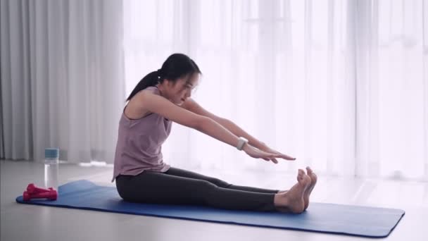 Una mujer asiática vestida con ropa de entrenamiento estira graciosamente sus músculos usando yoga o haciendo ejercicio posa sobre una alfombra en su sala de estar en casa. Ideal para la promoción de fitness o yoga. Imágenes de alta calidad 4k - Imágenes, Vídeo
