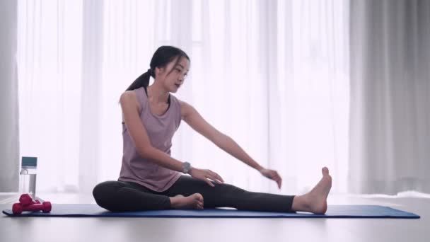 Een Aziatische vrouw in workout kleding strekt sierlijk haar spieren met behulp van yoga of oefening poseert op een mat in haar huis zitkamer. Ideaal voor fitness of yoga promotie. Hoge kwaliteit 4k beeldmateriaal - Video