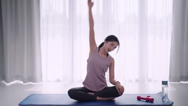 Una mujer asiática vestida con ropa de entrenamiento estira graciosamente sus músculos usando yoga o haciendo ejercicio posa sobre una alfombra en su sala de estar en casa. Ideal para la promoción de fitness o yoga. Imágenes de alta calidad 4k - Imágenes, Vídeo