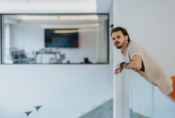 A thoughtful man with a beard leans on a glass partition in a modern office space, looking into the distance. Blurred office environment in the background evokes a sense of contemplation and focus. - Photo, Image