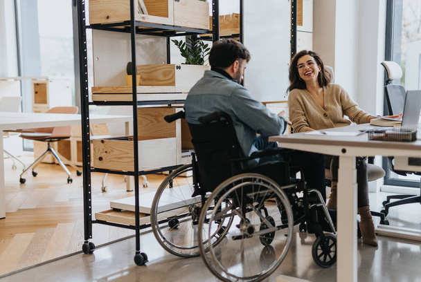 Úspěšná žena diskutuje o práci se svým hendikepovaným kolegou na invalidním vozíku v moderní kreativní kanceláři. - Fotografie, Obrázek