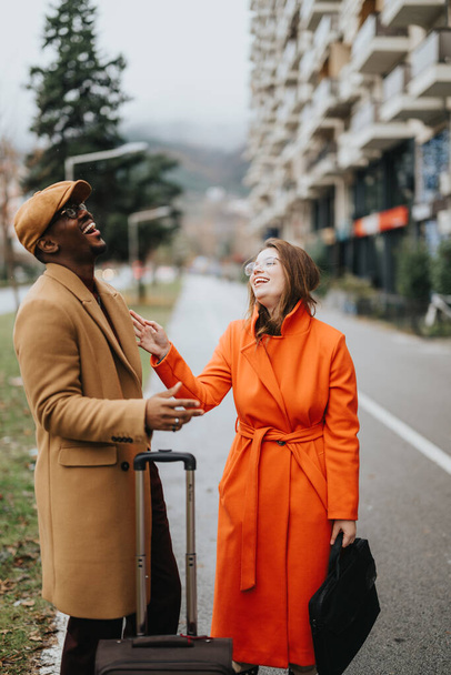 A fashionable man and woman laughing together on a city road, sharing a heartwarming moment with a suitcase suggesting travel or arrival. - Photo, Image