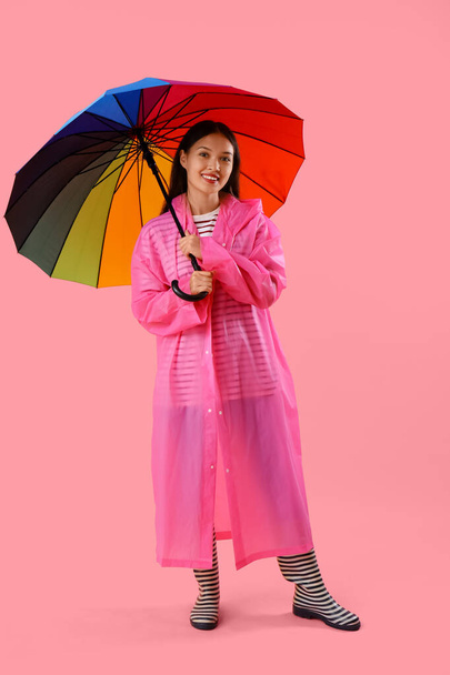 Mooie jonge Aziatische vrouw in regenjas met paraplu op roze achtergrond - Foto, afbeelding