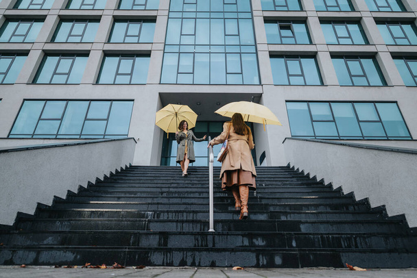 Sarı şemsiyeli sıradan iş kadınları modern bir ofis binasından çıkıyorlar bulutlu bir günde merdivenlerden iniyorlar şehir yaşamını ve dostluğunu sergiliyorlar.. - Fotoğraf, Görsel