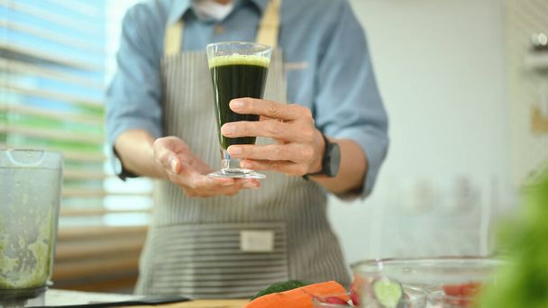 Hombre mayor saludable sosteniendo un vaso de batido de verduras verdes cerca de los ingredientes en la cocina. - Foto, imagen