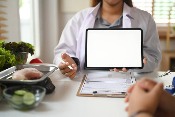 Nutricionista profesional en bata blanca mostrando tableta digital y dando consulta al paciente en clínica. - Foto, imagen