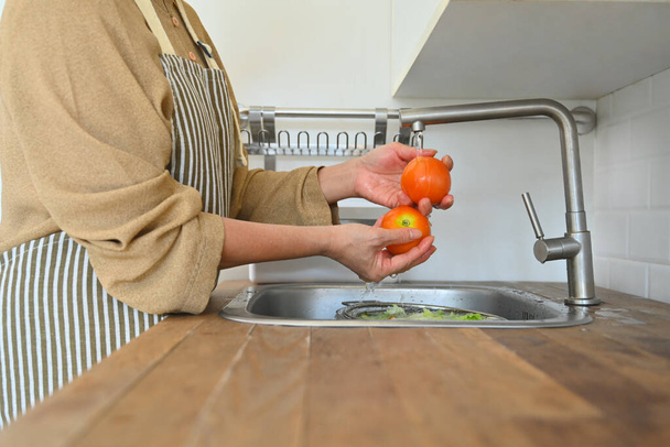 Καλλιεργούνται πυροβόλησε ανώτερος νοικοκυρά στην ποδιά πλύσιμο φρέσκα λαχανικά για σαλάτα στην κουζίνα. - Φωτογραφία, εικόνα
