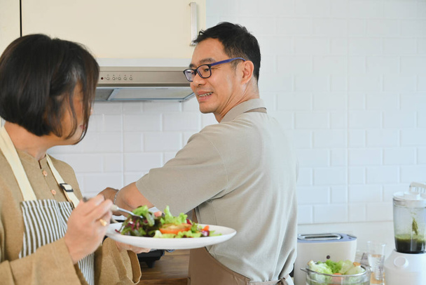 Привлекательная пожилая пара наслаждается здоровым обедом в уютном кухонном интерьере. Концепция ухода на пенсию и здорового питания. - Фото, изображение