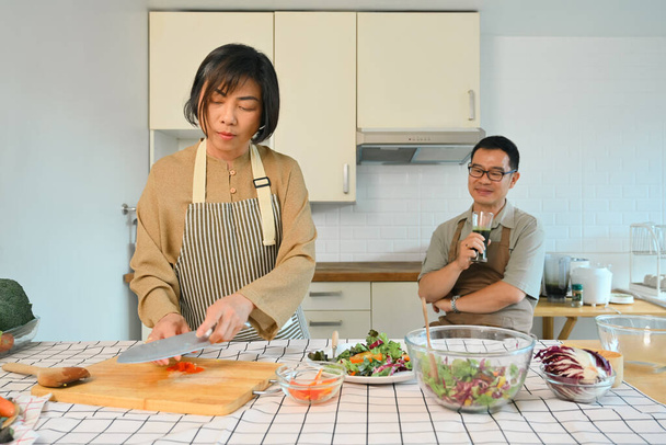 Heureuse femme d'âge moyen faisant salade de légumes dans la cuisine, passer du temps avec son mari à la maison. - Photo, image