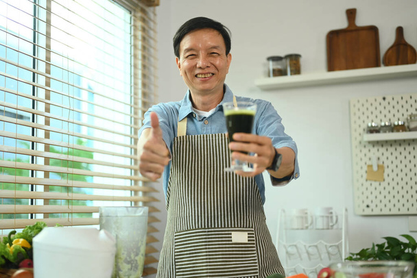 Fröhlicher Mann mittleren Alters hält ein Glas frischen Gemüse-Smoothie in der Hand und zeigt Daumen hoch. - Foto, Bild