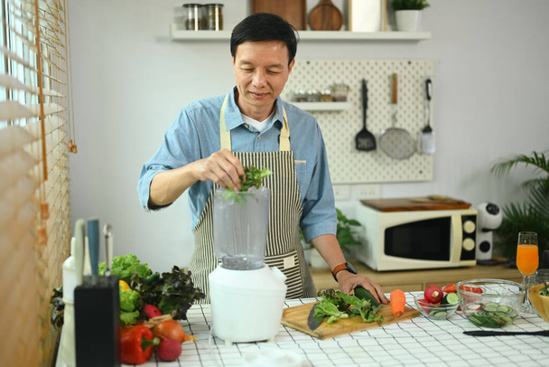 Happy middle age man making vegetable smoothies with blender in kitchen. Healthy lifestyle concept. - Photo, Image