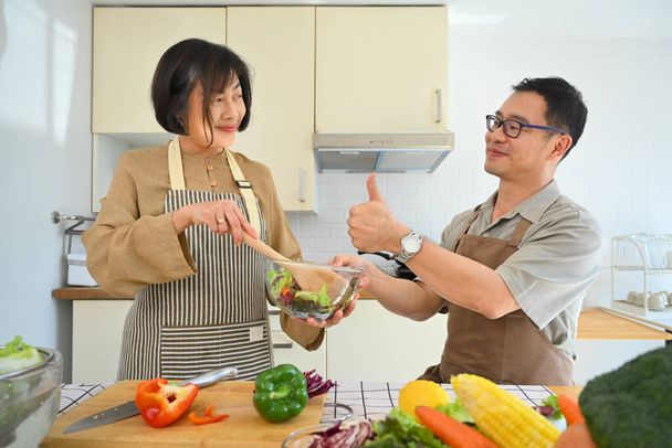 Glückliches Ehepaar mittleren Alters kocht gesunde vegetarische Mahlzeit und unterhält sich in der Küche. - Foto, Bild