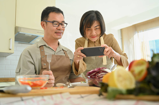 Happy senior woman taking photo of vegan meal with smartphone while cooking with her husband in kitchen - Photo, Image
