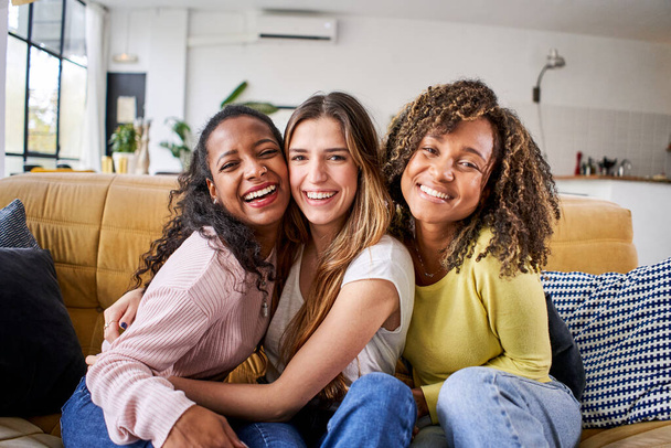 Porträt von drei attraktiven weiblichen Mitbewohnerinnen, die lächelnd in die Kamera schauen. Gruppe von netten, fröhlichen Mädchen, die sich umarmen, wenn sie zu Hause auf der Couch sitzen. Glückliche Freunde posieren für ein Foto im Haus. Generation z und Miete. - Foto, Bild