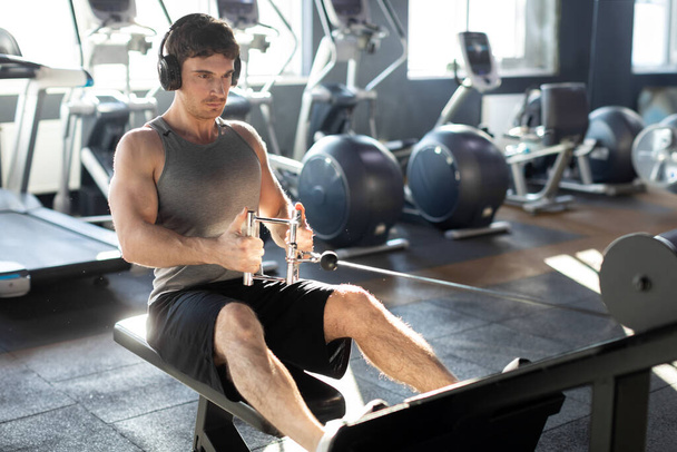 Молодой мускулистый мужчина фокусируется на тренировке на кабельном, носит наушники и слушает музыку, на фоне тяжелых пластин и оборудования - Фото, изображение