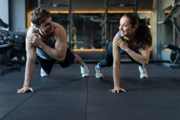 Young athlete couple making one hand plank exercise together, looking and smiling to each other, training at modern gym interior - Photo, Image