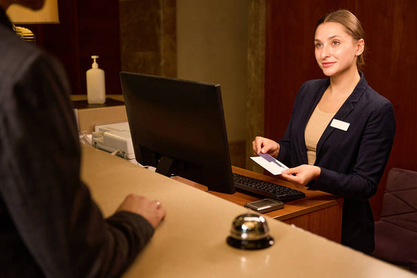 Amichevole donna receptionist mettendo keycard in busta di carta mentre in piedi alla reception e guardando il visitatore - Foto, immagini
