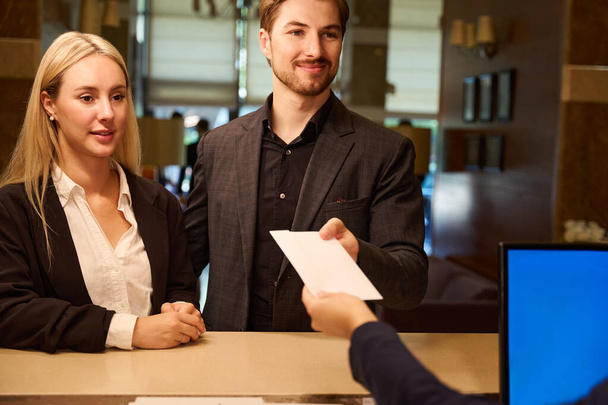 Młody mężczyzna i kobieta stojący w recepcji w hotelu podczas gdy mężczyzna bierze papierową kopertę od administratora - Zdjęcie, obraz