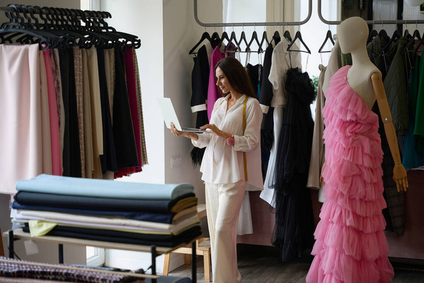 Pregnant woman with a laptop stands in a design studio, next to a mannequin in a pink evening dress - Photo, Image
