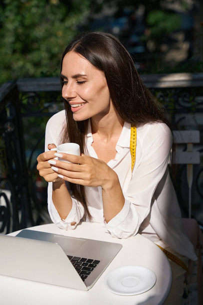 Νεαρή γυναίκα απολαμβάνει ένα φλιτζάνι καφέ σε ένα γοητευτικό μπαλκόνι με μια μεζούρα γύρω από το λαιμό της - Φωτογραφία, εικόνα