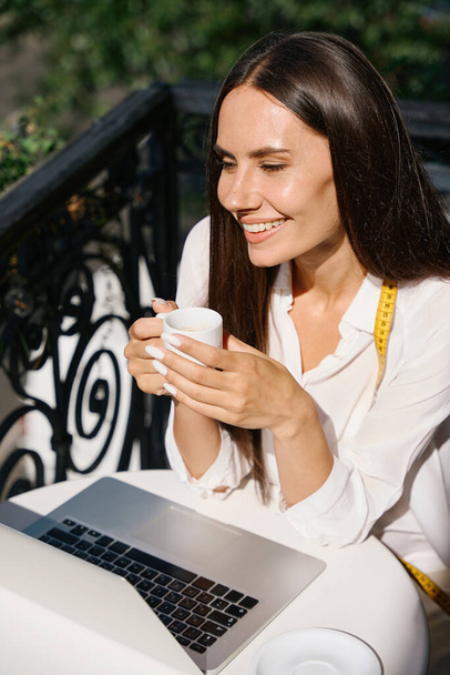 Mujer sonriente disfrutando de una taza de café en un encantador balcón con una cinta métrica alrededor de su cuello - Foto, imagen