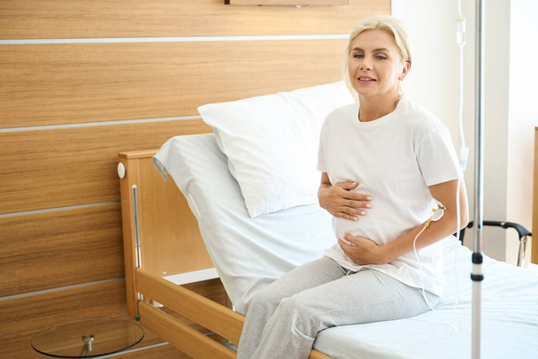 Przemyślana dorosła Europejka w ciąży, siedząca na łóżku medycznym i obejmująca brzuch w szpitalu położniczym - Zdjęcie, obraz