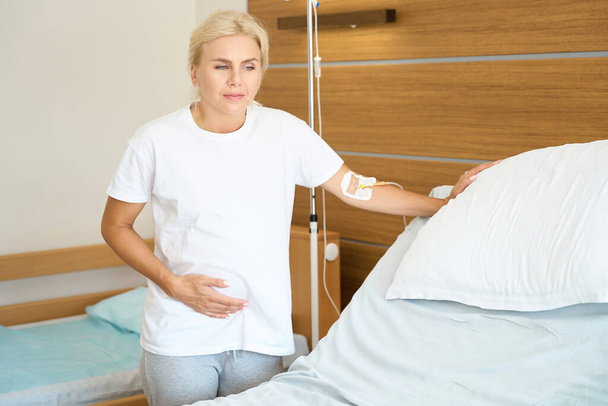 Dorosła Europejka w ciąży, stojąca obok łóżka medycznego i dotykająca brzucha w szpitalu położniczym - Zdjęcie, obraz