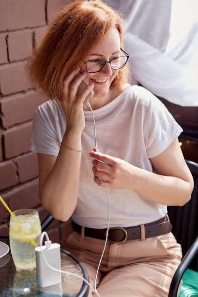 Flessibilità freelance: una donna dai capelli rossi carica il telefono con una powerbank mentre lavora da remoto. - Foto, immagini