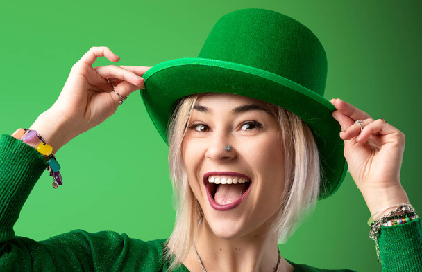 St. Patrick 's Dag. Joyful meisje in een kabouter hoed viert St. Patrick 's Day in een groene trui. Feestelijke geest gevangen tegen een levendige groene achtergrond - Foto, afbeelding