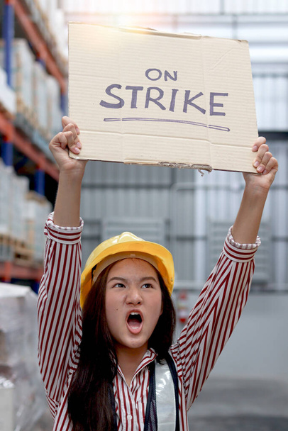 Wütend unzufriedene asiatische Arbeiterinnen mit Warnweste und Helm, die ein Schild mit einem Streikbanner am Frachtlogistiklager halten und hochhalten. Streikende Arbeiter protestieren am Arbeitsplatz. - Foto, Bild