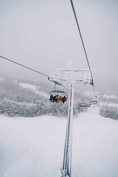 Turistit värikkäissä hiihtopuvuissa ratsastaa tuolihissillä yli luminen metsä ylämäkeen. Laadukas kuva - Valokuva, kuva