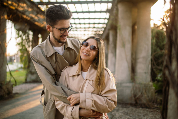 Portrait of a girl and a guy posing against the backdrop of an arch in the garden at sunset in spring. Family photo of a stylish couple in beige trench coats - Photo, Image