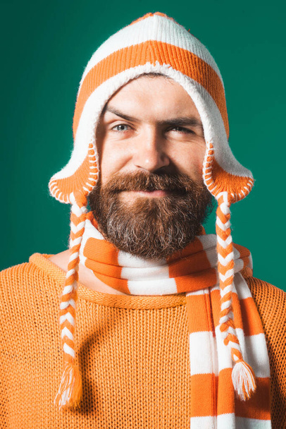 Крупный план портрета привлекательного мужчины с бородой и усами в оранжевом свитере, смешной шляпе и полосатом шарфе. Красивый бородатый мужчина в теплой одежде. Осенне-зимняя одежда. Повседневный взгляд. Мода и стиль - Фото, изображение