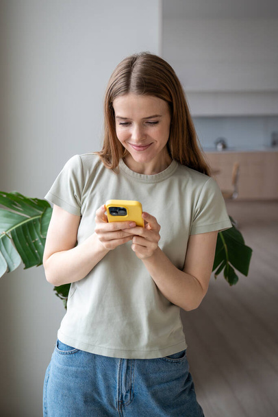 Tyytyväinen nainen lukee viestejä matkapuhelimella kotona ikkunan lähellä. Hymyilevä tyttö juttelee älypuhelimessa ystävien kanssa. Positiivinen nuori nainen nauttia rentouttava gadget. Sosiaalisen median riippuvuus.  - Valokuva, kuva