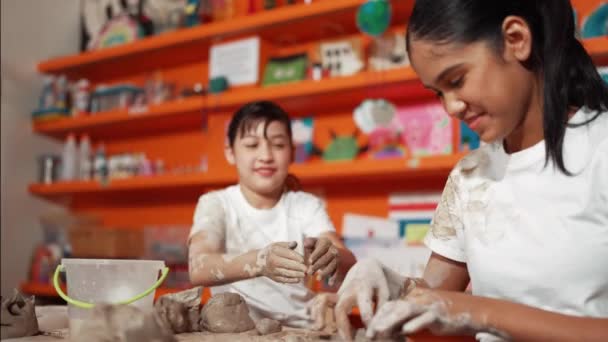 アジアの子供は陶器のクラスで白人少女のシャツに粘土を置きました. アートレッスンでスマートな友人と粘土を再生する混合レースと幸せな学生. 教育について。 イスラーム主義 - 映像、動画