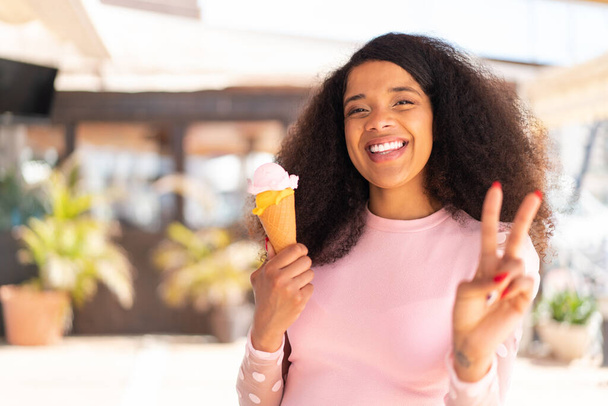 Jonge Afro-Amerikaanse vrouw met een cornet ijs in de buitenlucht glimlachen en tonen overwinning teken - Foto, afbeelding