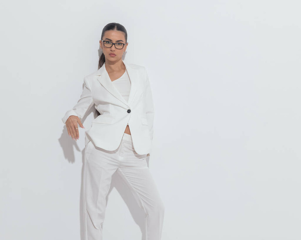 cool elegant girl with glasses holding hands in pockets and posing in a confident way in front of grey background - Photo, Image