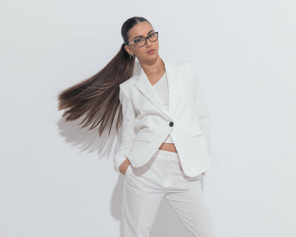 elegant businesswoman with glasses flipping hair and holding hands in pockets while posing in front of grey background - Photo, Image