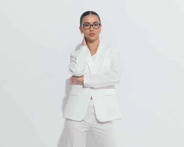 привлекательная молодая женщина в очках в белом костюме держит руку на шее и складывает руки на белом фоне - Фото, изображение