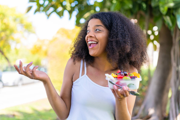 Junge Afroamerikanerin hält mit überraschendem Gesichtsausdruck eine Schale mit Früchten in der Hand - Foto, Bild