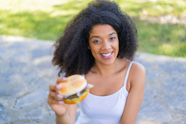 Junge Afroamerikanerin hält draußen mit glücklichem Gesichtsausdruck einen Burger in der Hand - Foto, Bild