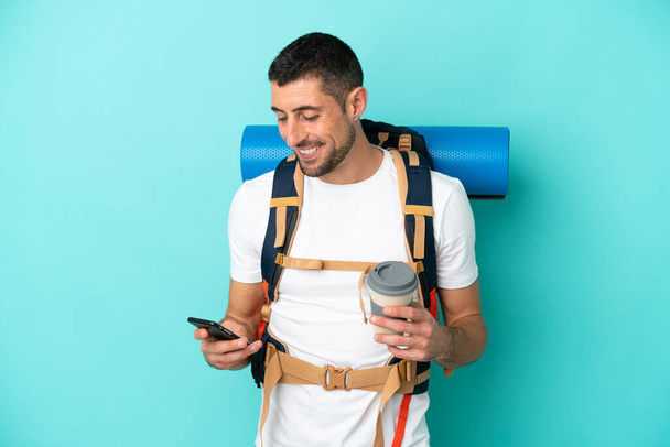 Молодий альпініст кавказький чоловік з великим рюкзаком ізольований на синьому фоні, тримаючи каву, щоб забрати і мобільний
 - Фото, зображення