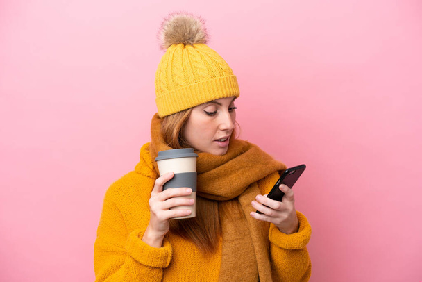 Fiatal vörös hajú nő visel téli kabát elszigetelt rózsaszín háttér gazdaság kávé, hogy vigye el, és egy mobil - Fotó, kép