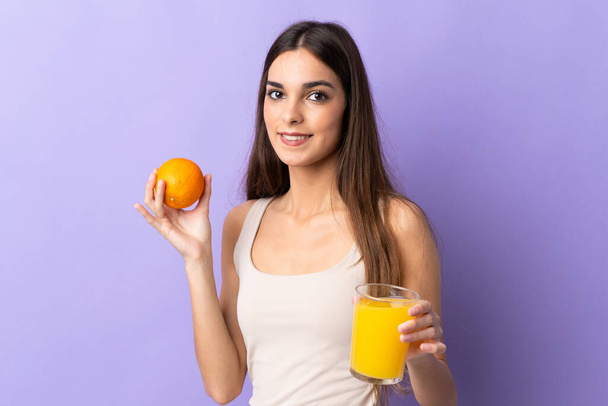 Mujer caucásica joven aislada sobre fondo púrpura sosteniendo una naranja y un jugo de naranja
 - Foto, imagen