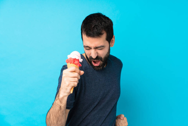Jeune homme avec une crème glacée cornet sur fond bleu isolé célébrant une victoire - Photo, image