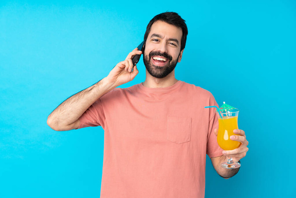 Junger Mann hält einen Cocktail vor isoliertem blauen Hintergrund und hält ein Gespräch mit dem Handy mit jemandem - Foto, Bild