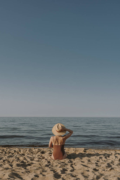 Beautiful woman in straw hat and swimsuit sitting on sand at beach and watching at sea and sky. Aesthetic summer fashion concept - Photo, Image