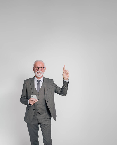 Usmívající se starší muž podnikatel s mobilním telefonem ukazuje nahoru a marketing na bílém pozadí - Fotografie, Obrázek